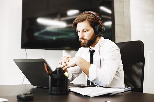 Cuffie smart working: uomo in camicia e cravatta in ufficio in videochiamata a computer con le cuffie